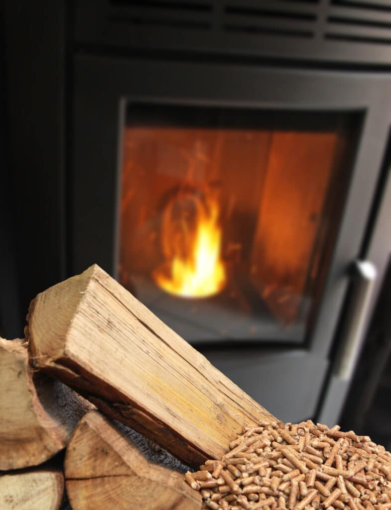 Bûches et granulés de bois empilés, avec un poêle à bois noir et ses flammes ardentes en arrière-plan.